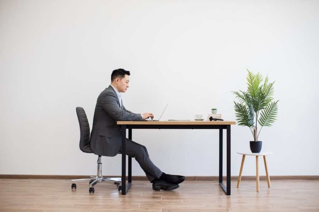 Asian businessman in suit sitting at desk working on laptop. Modern minimalist office setting with wooden floor, white wall, and a potted plant.
